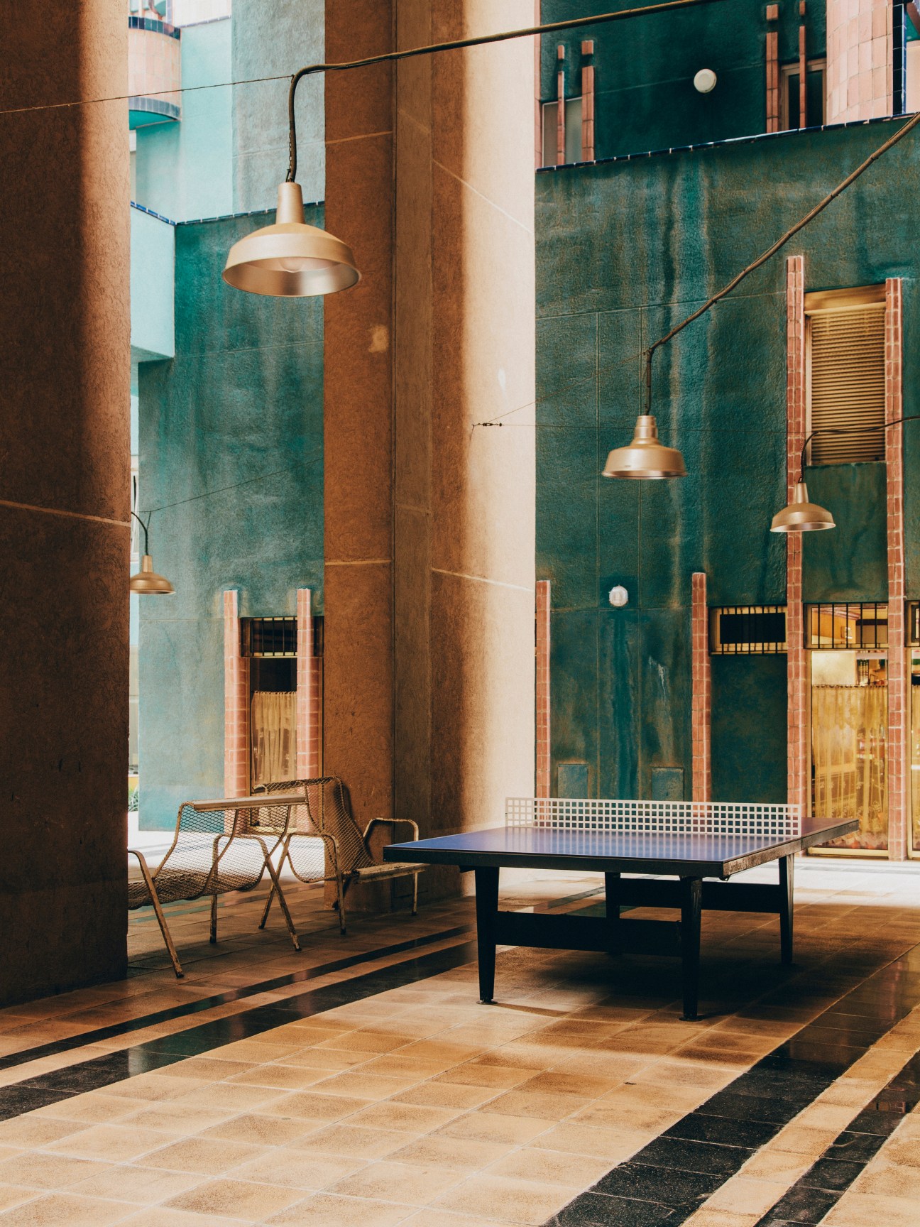 Communal ping pong table in Walden 7's ground floor courtyard with turquoise walls, brass pendant lights, and wooden chairs.
