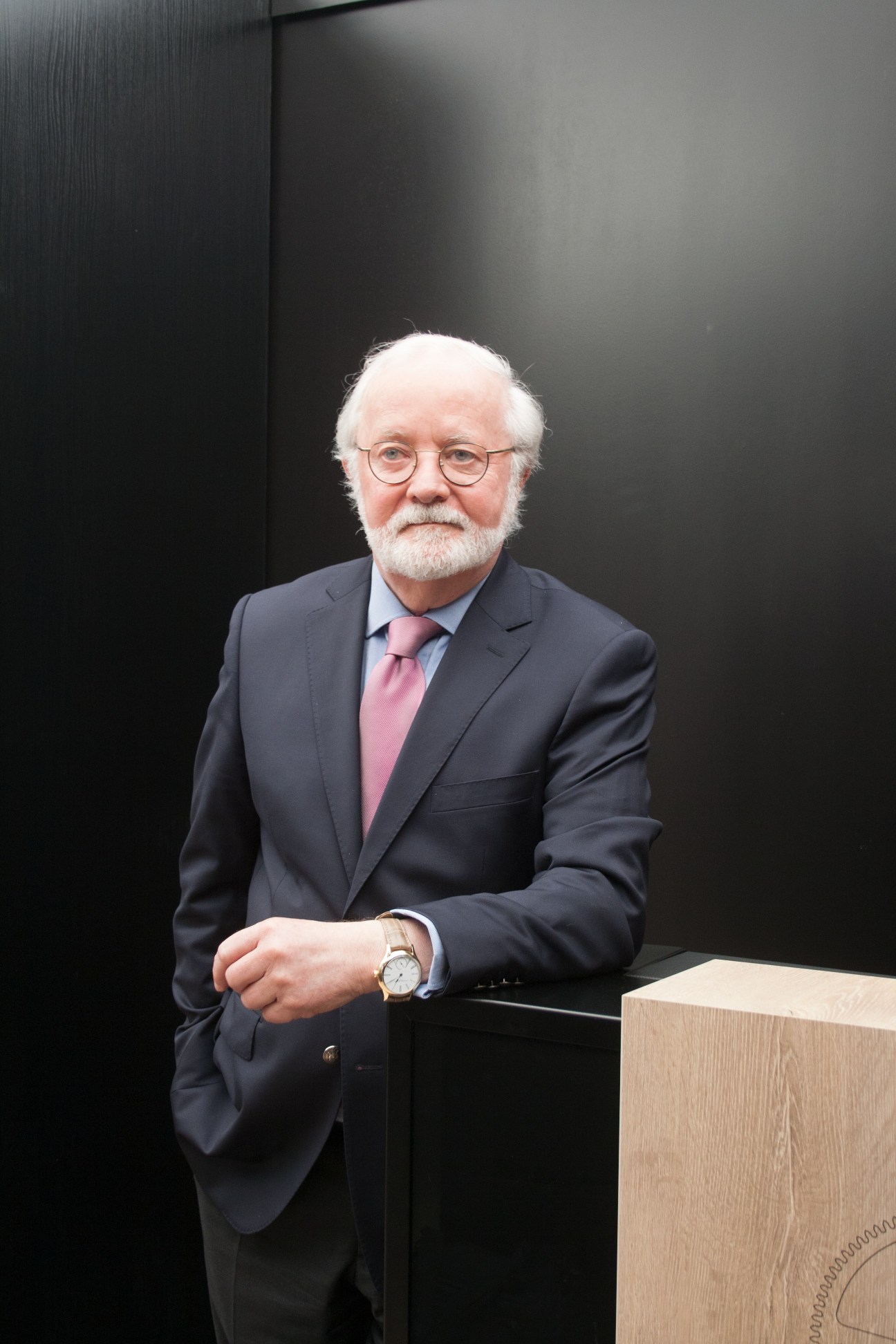 Elderly man with white beard and glasses wearing dark suit and pink tie, displaying watch on wrist.