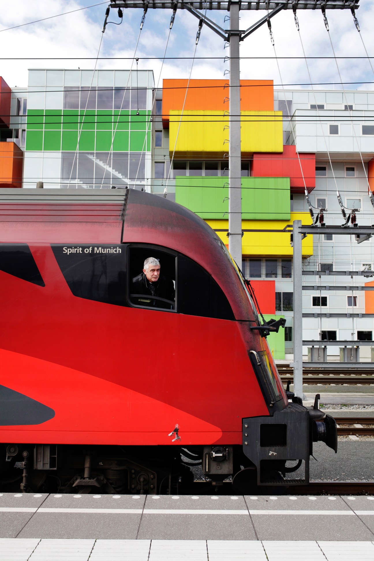 Red ÖBB Railjet train with driver visible in cabin window, colorful building facade behind.