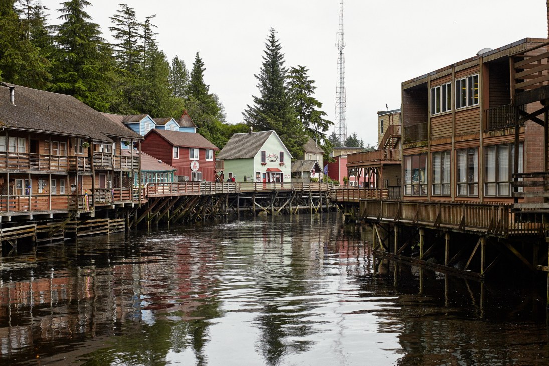 Historic wooden buildings line a narrow waterway in Ketchikan, Alaska, with coniferous trees and a communications tower in the background.