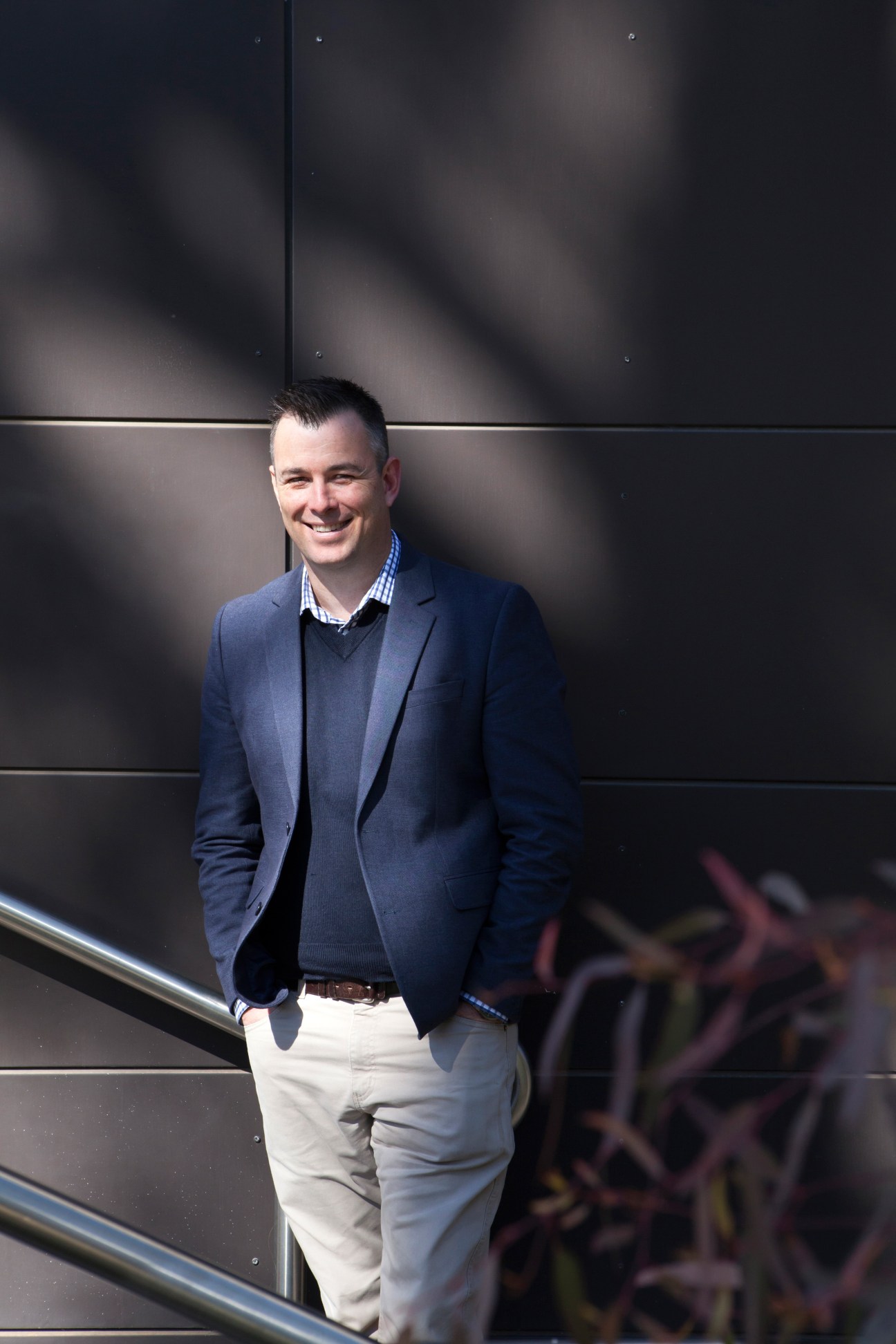 Senior fellow Dr Peter Dean stands smiling against a dark textured wall.