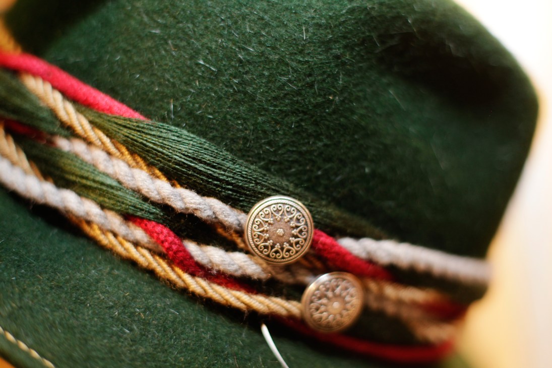 Close-up of a green felt alpine hat with decorative metal buttons and red, gold and white braided trim by Bittner.
