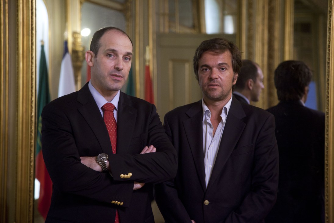 Manuel Lapão and António Ilharco stand in an ornate palace corridor with flags in the background.