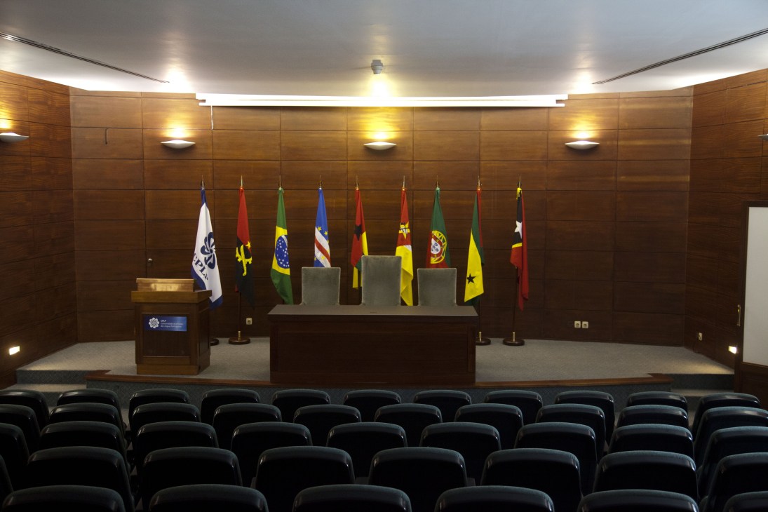 CPLP member nation flags arranged alphabetically in a wood-paneled conference hall with podium and seating.