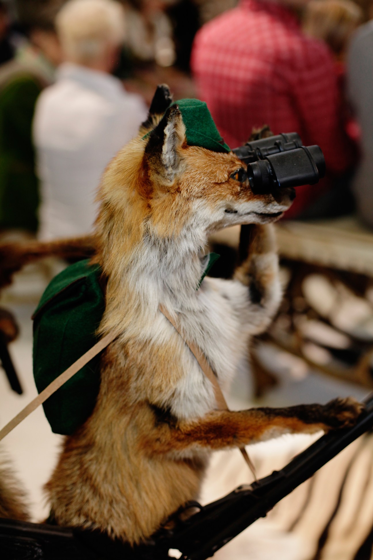 Taxidermied deer wearing a green alpine hat and binoculars at a trade fair display.