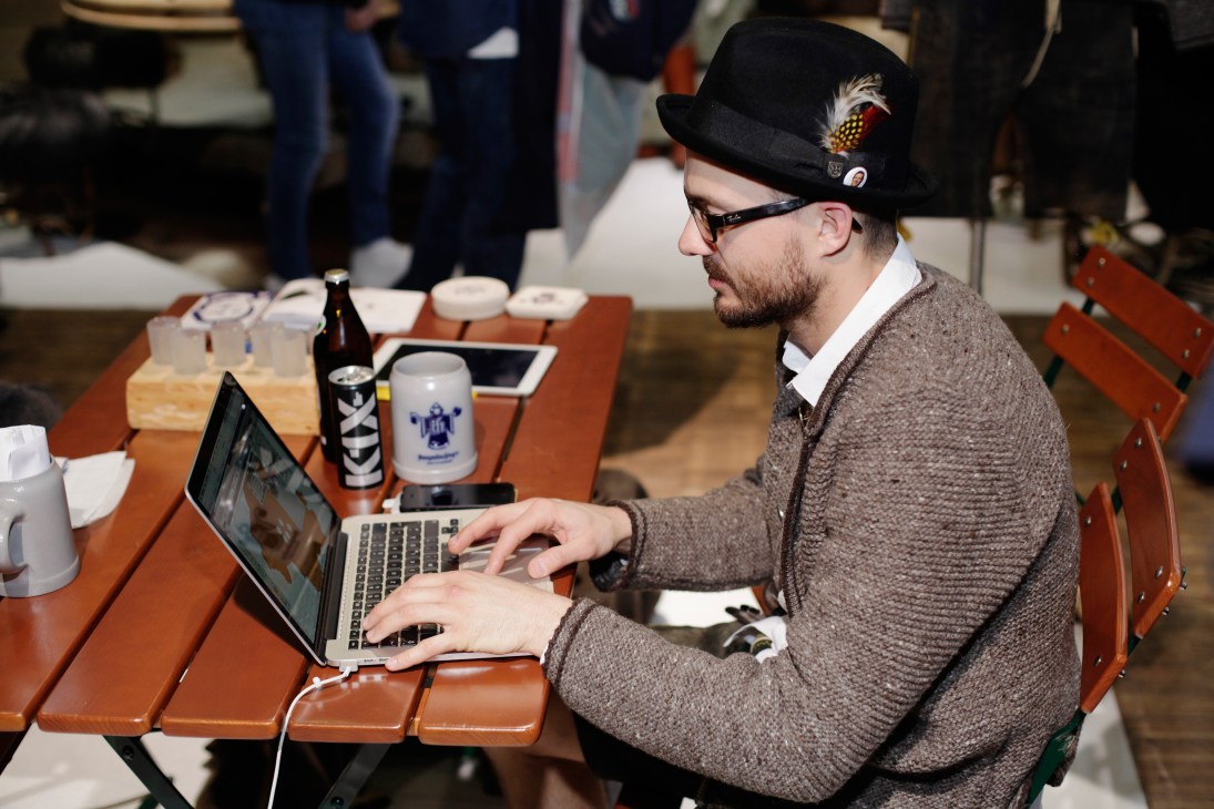Man in traditional Alpine hat and brown cardigan working on laptop at fair table
