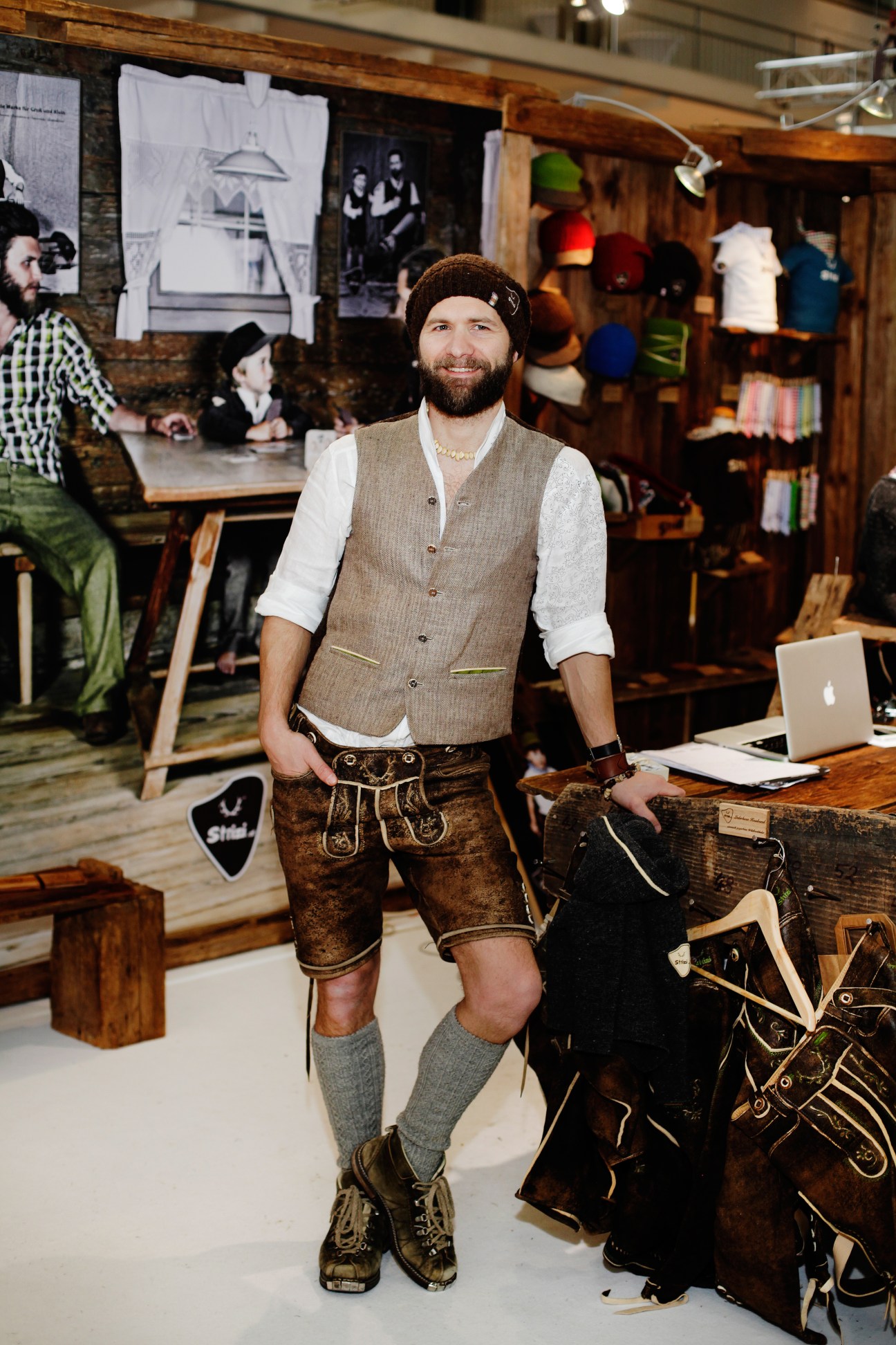Bernhard Stadler, owner of Strizi design, wearing traditional alpine clothing including a beige waistcoat, leather shorts, and knitted beanie in his shop.