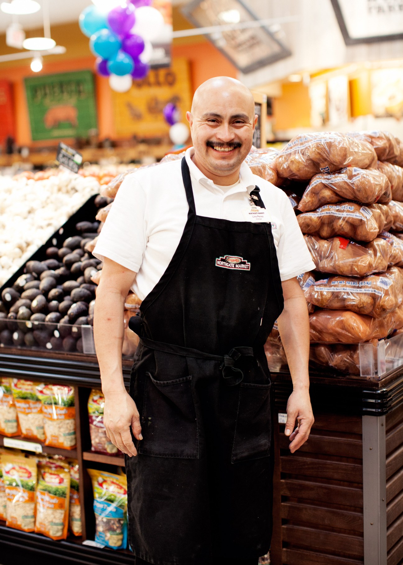 Lino Perez, a produce worker at Northgate Markets in Los Angeles, smiling in front of stacked packages of food in a colorful supermarket.