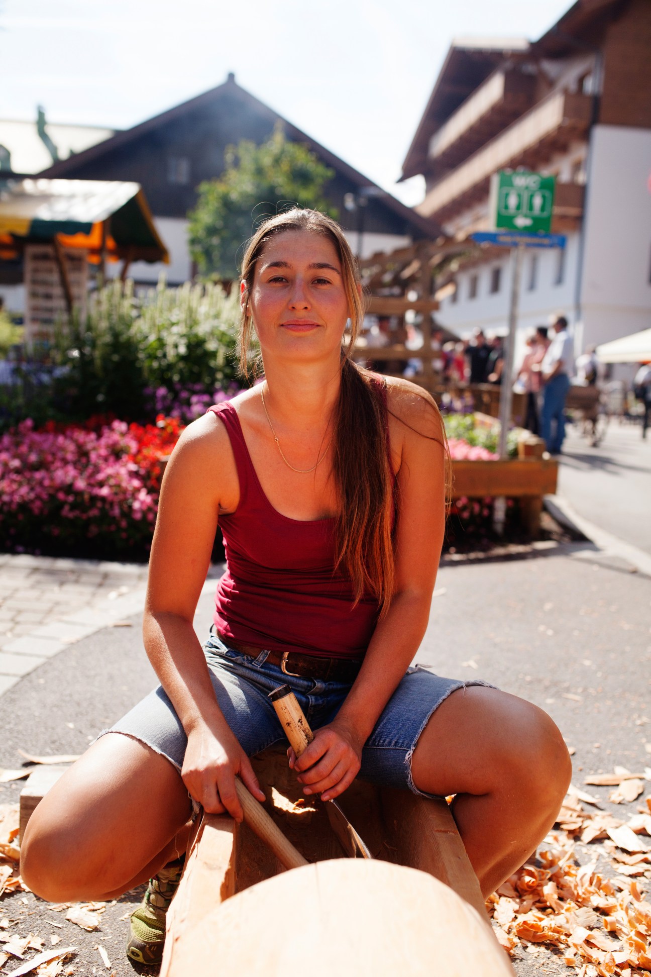 Sandra Neuner sits cross-legged outdoors holding wood carving tools with traditional Alpine buildings in background
