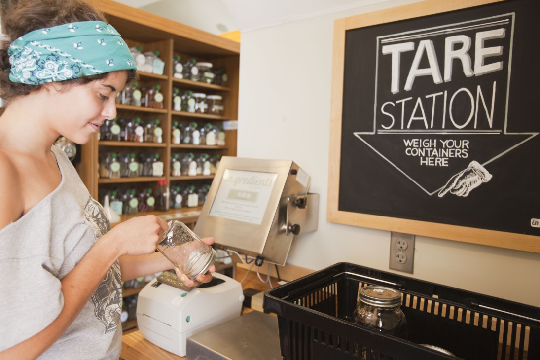 Woman wearing a blue headband weighing a glass jar at In.gredients waste-free grocery store tare station.