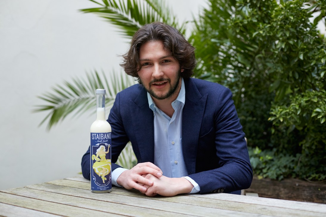 Cesco Amodio poses with a bottle of Staibano lemon liqueur on a wooden table with tropical plants behind him.