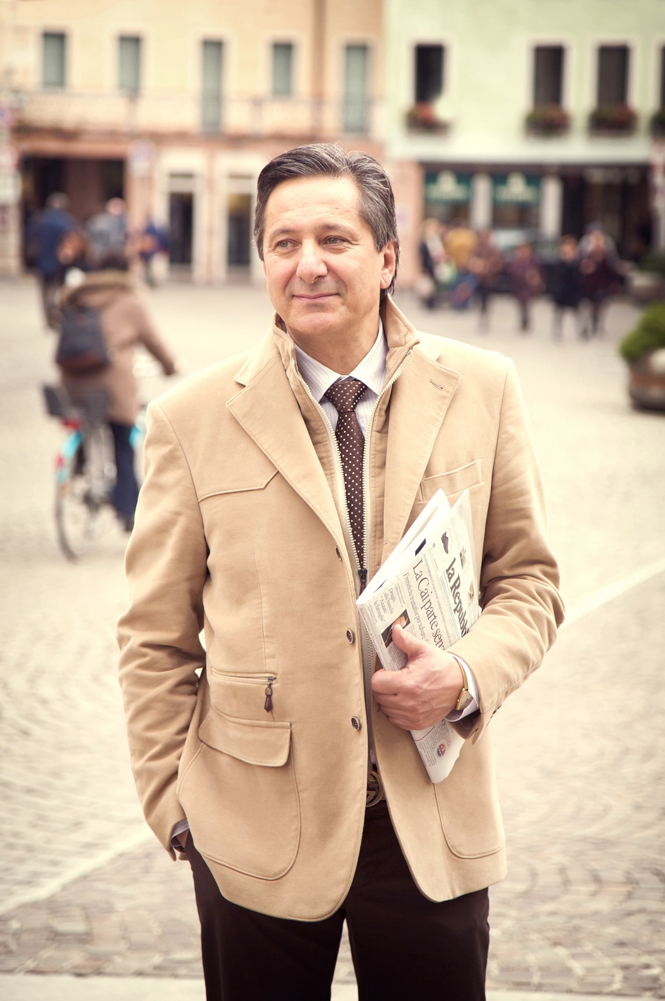Psychologist Renzo Vettorazzi stands in a town square holding a newspaper, wearing a beige blazer and brown tie.