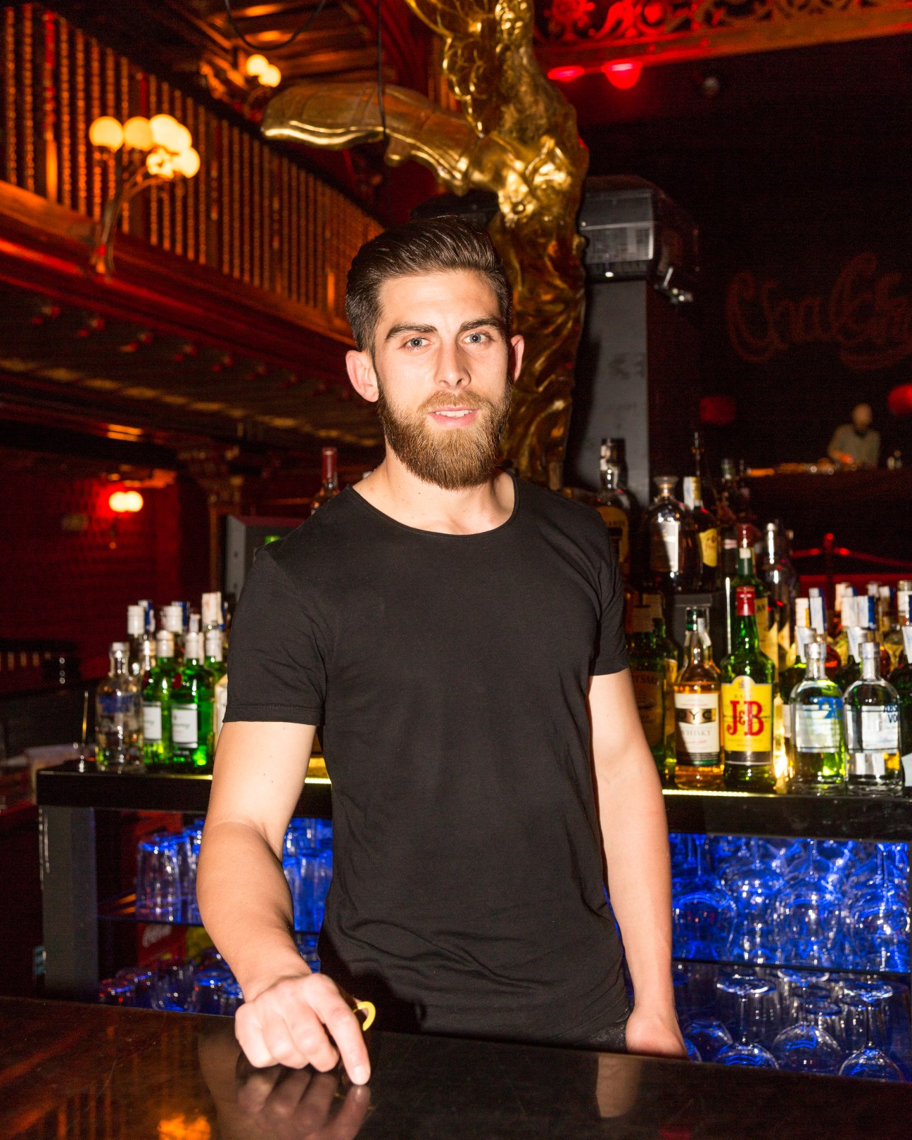 Bartender in black t-shirt standing at a bar with bottles and glasses, surrounded by red and gold lighting.