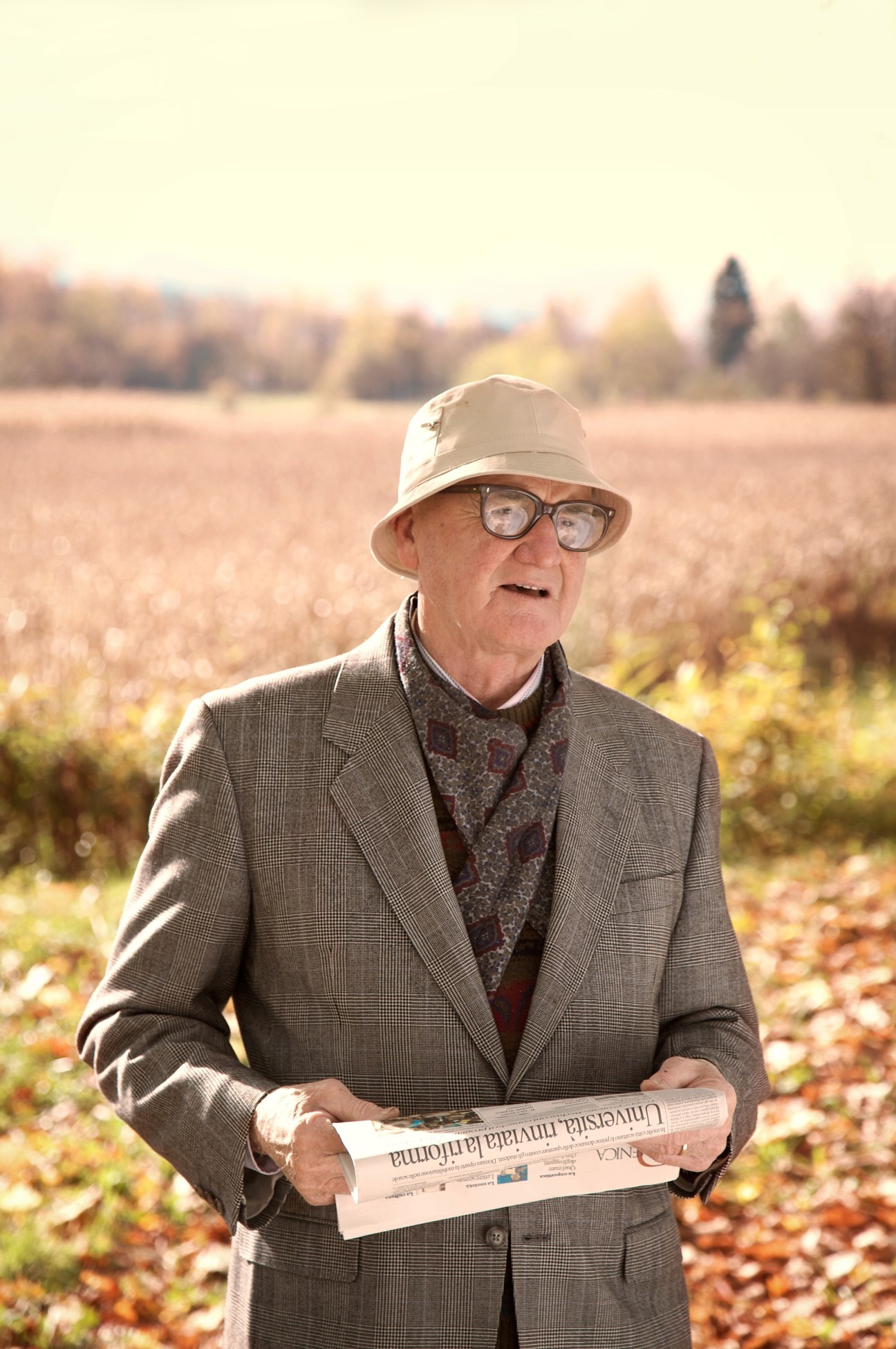 Retired electrician Tullio Vallata stands in a field holding a newspaper, wearing glasses and a hat.