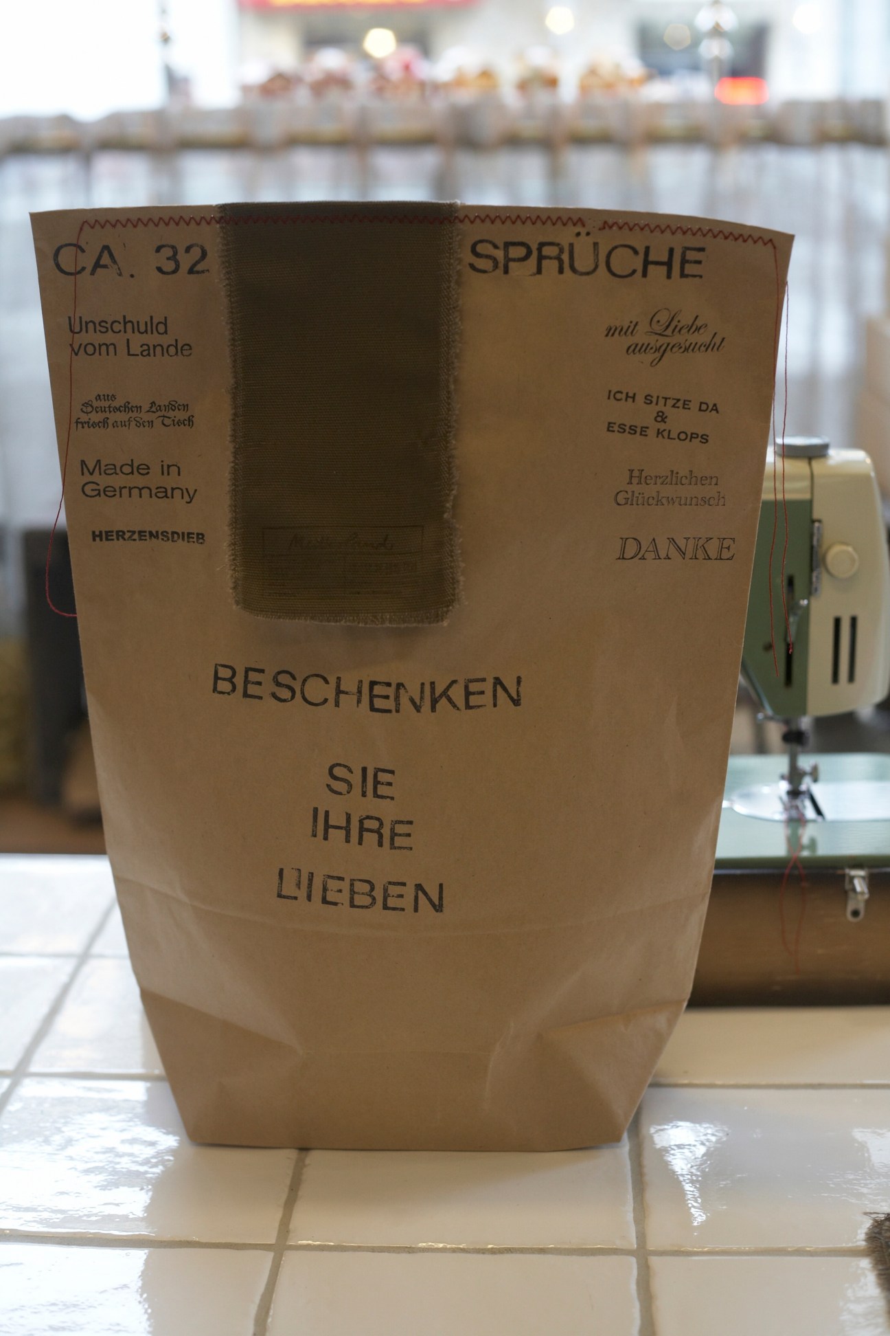 Brown paper gift bag with German text sewn closed with red thread on a sewing machine visible in background.