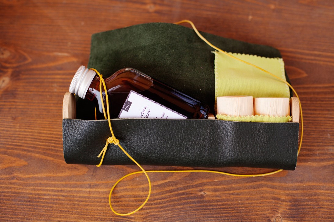 Black leather tool roll containing glass bottle, small jars, and yellow polishing cloth on wooden surface