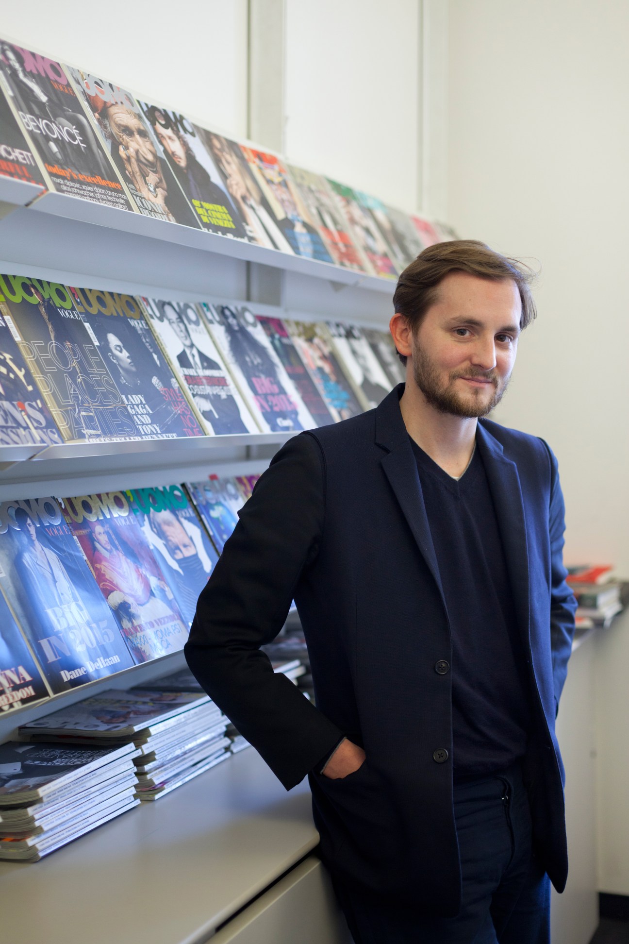 Alan Prada, managing editor of L'Uomo Vogue, stands in front of shelves displaying magazine covers.