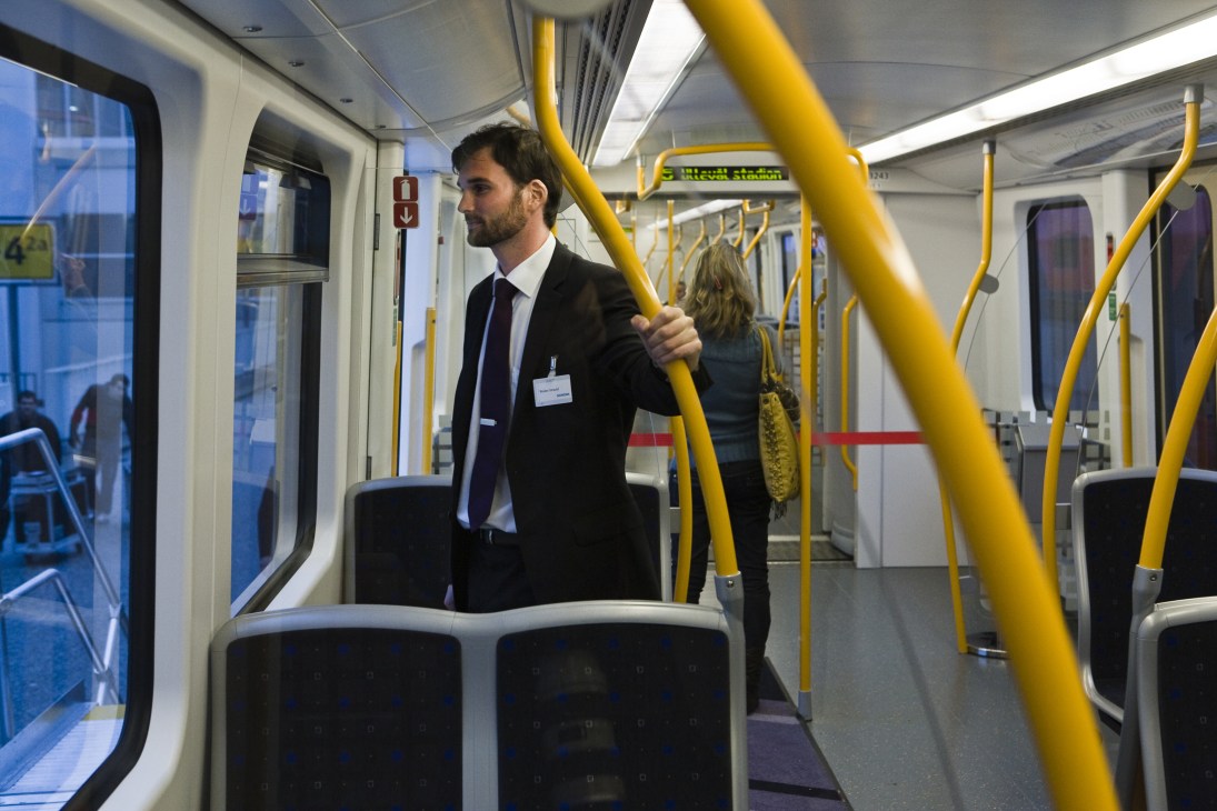 A man in a dark suit and tie stands inside a modern metro train car with yellow handrails and purple seats.