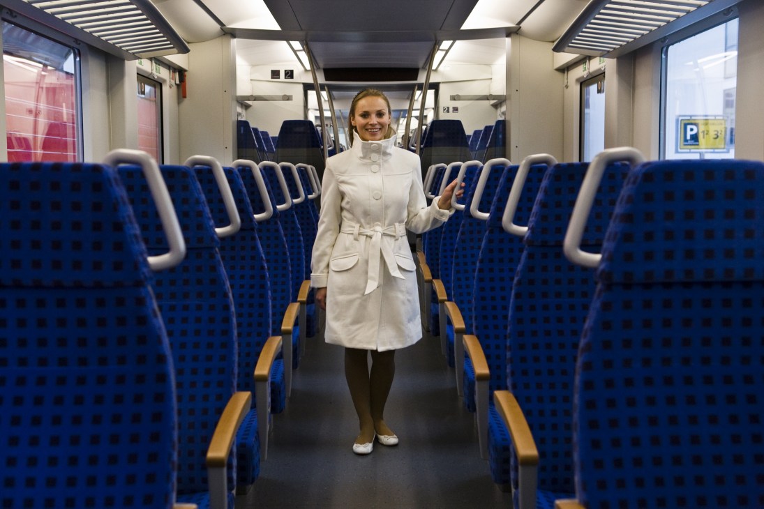 Train attendant in white coat stands in aisle of German DB Inter Regio-train by Alstom with blue seats.