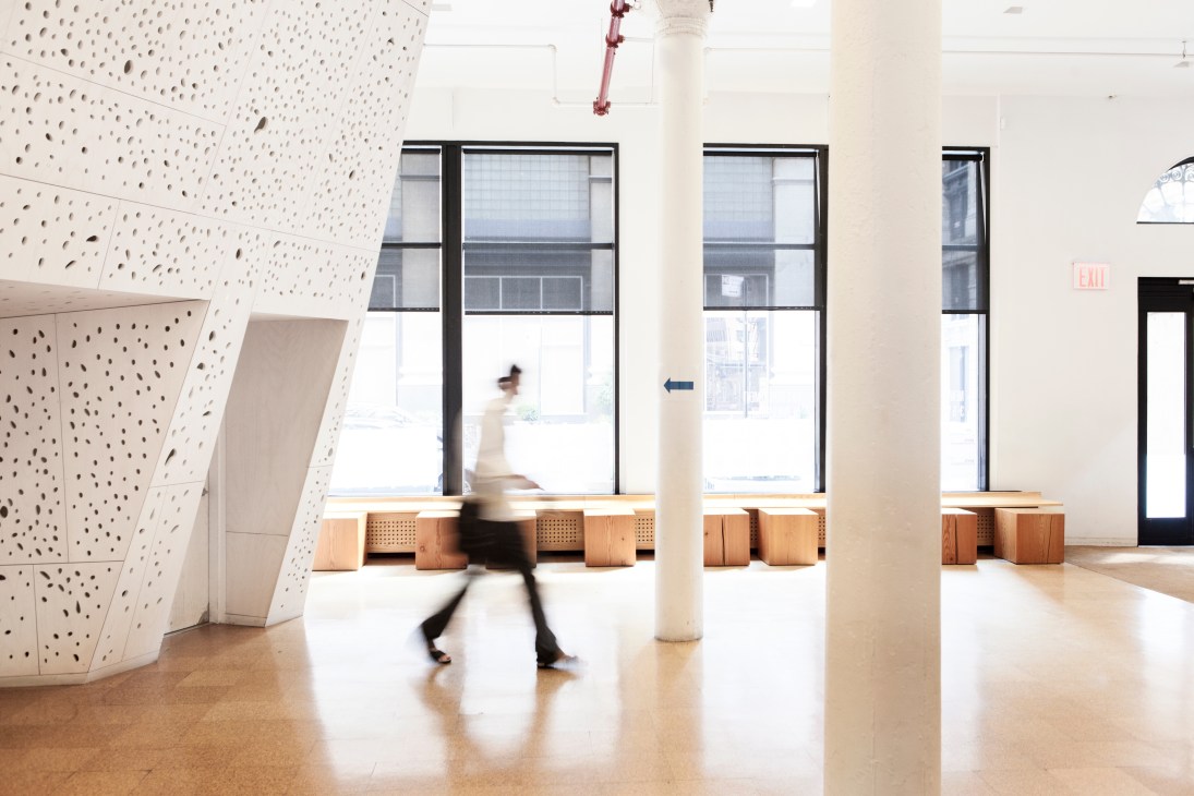 Bright modern lobby with perforated white walls, wooden seating, large windows, and a person walking through the space.