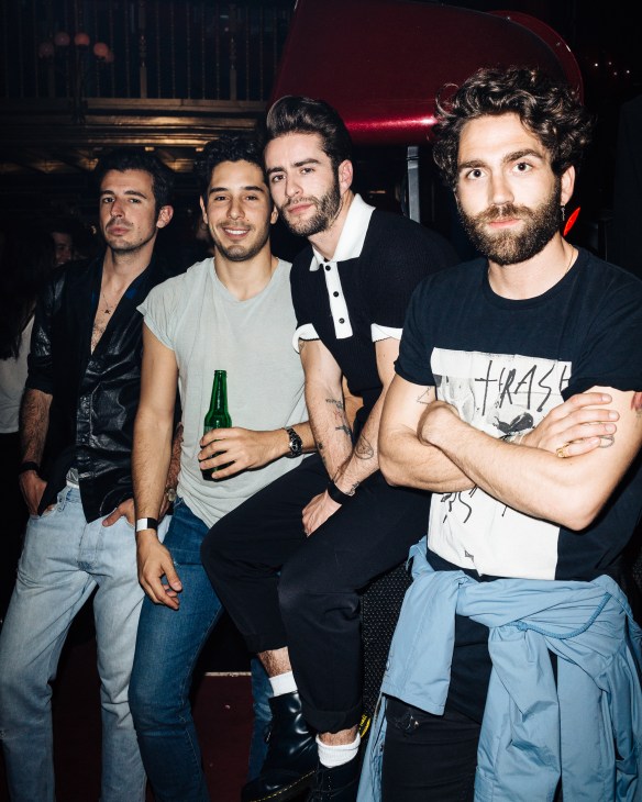 Four men posing together inside a nightclub with red lighting and velvet furnishings.