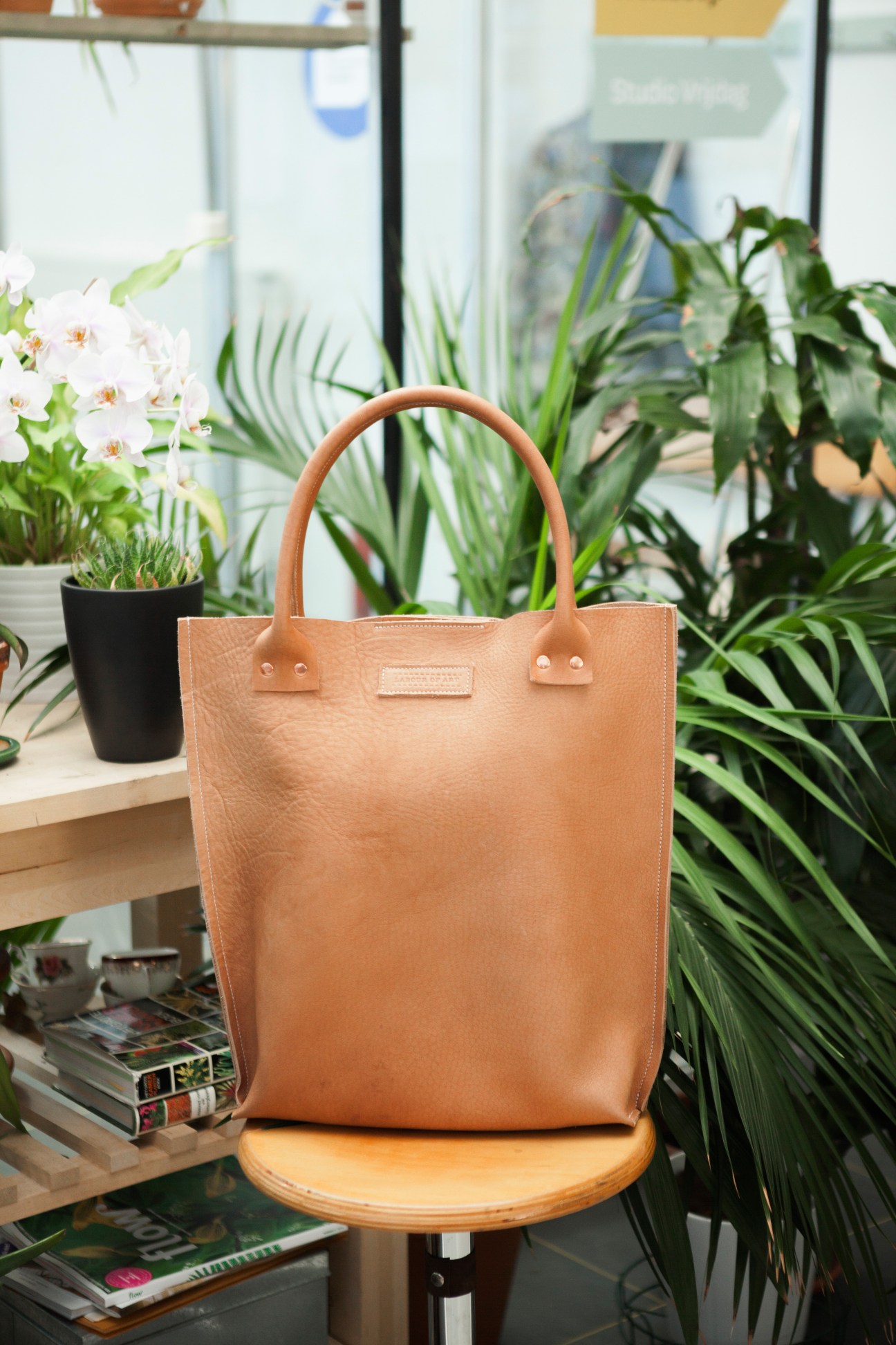 Tan leather tote bag displayed on wooden chair surrounded by potted plants and magazines