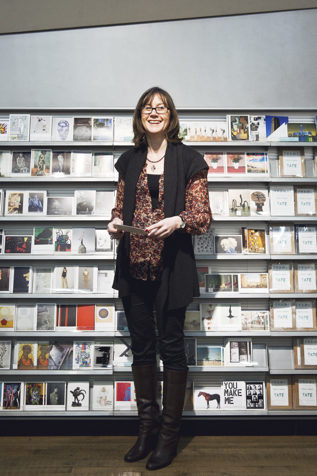 Rosey Blackmore, merchandise director at Tate Modern, stands smiling in front of shelves displaying postcards.