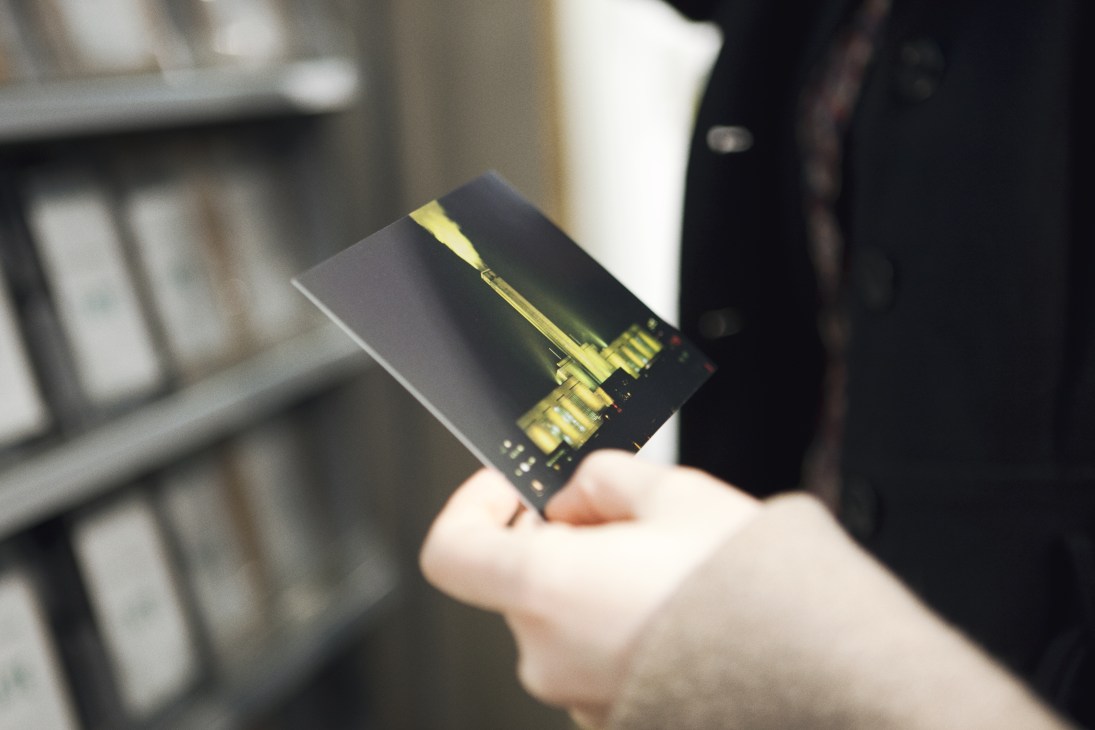 A hand holding a postcard featuring the illuminated Tate Modern building at night against a dark background.