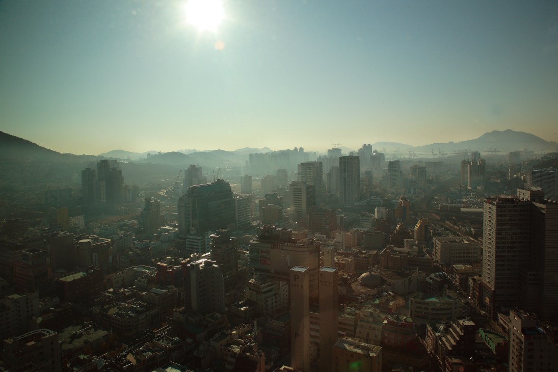 Aerial view of Busan cityscape with high-rise buildings and mountains in hazy distance.