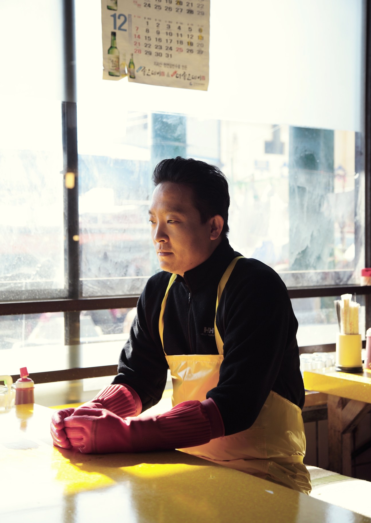 A worker at Busan's fish market wearing a yellow apron and red gloves, sitting at a table with a city skyline visible through the window behind him.