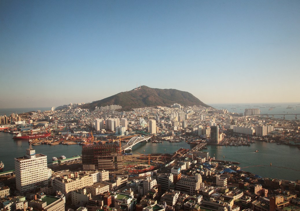 Aerial view of Busan's waterfront showing a mountain, dense urban buildings, harbor with ships and industrial facilities.