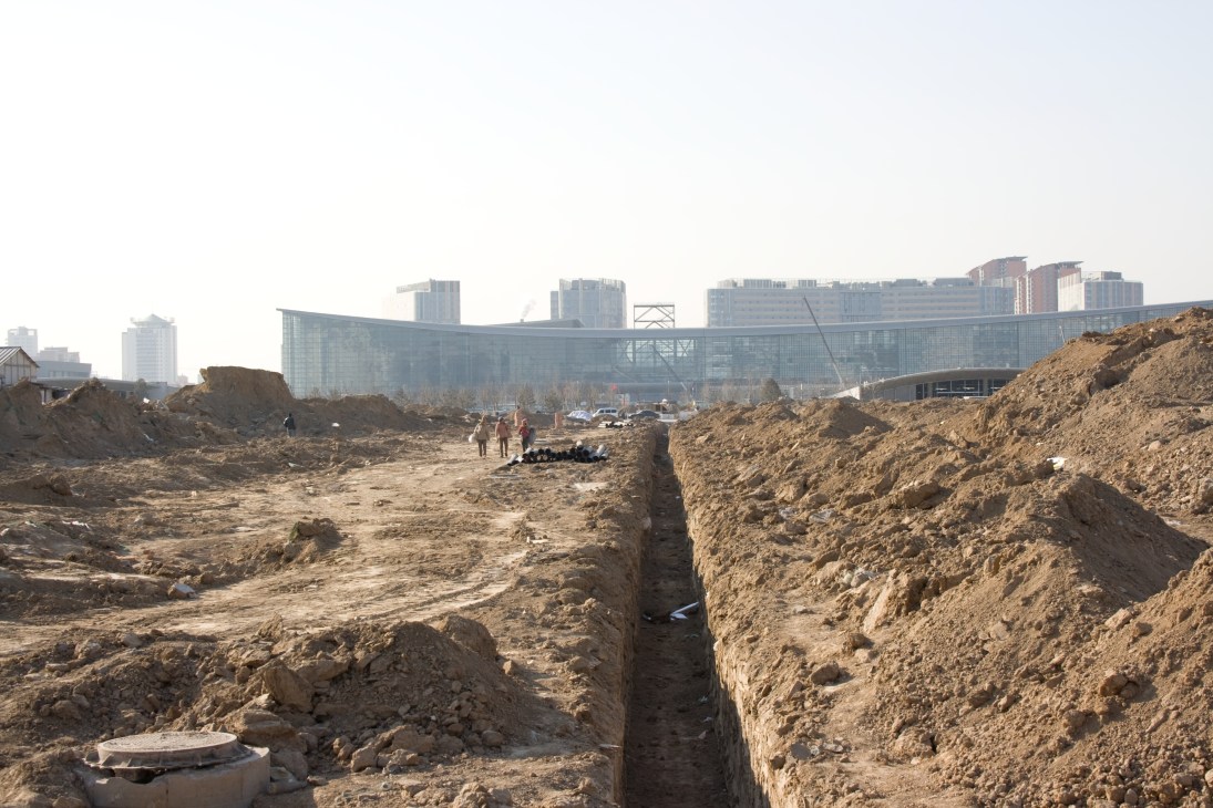 Construction site with excavated earth and trenches, National Convention Centre building visible in background among apartment towers.
