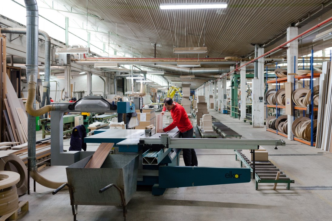 Workers operating machinery and processing wooden materials inside a large, industrial furniture factory with metal beams and storage racks.