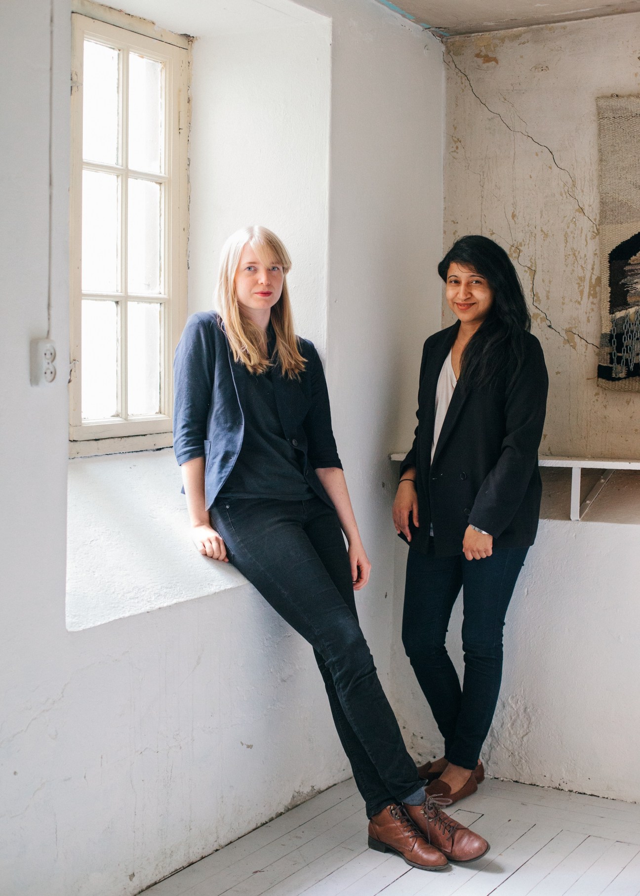 Two women pose in an industrial gallery space with white walls and a large window.