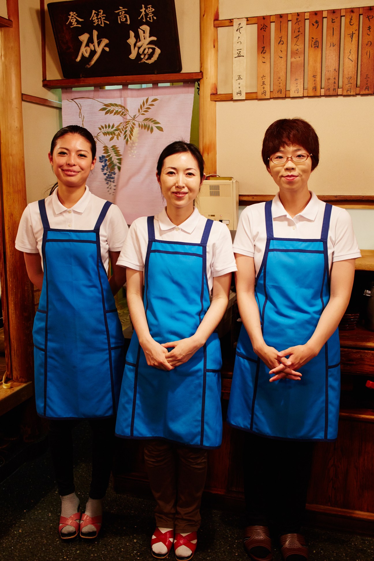 Three women in blue aprons and white shirts stand together in a traditional Japanese restaurant interior.