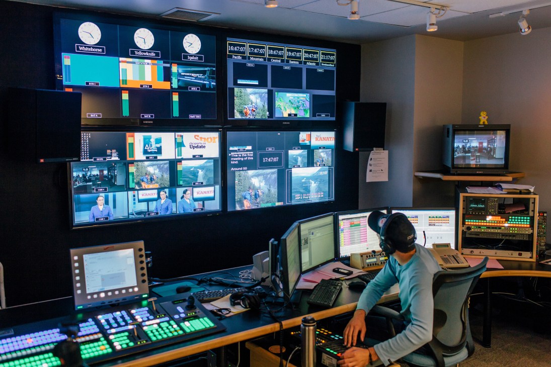 Technical director Allan Gofenko sits at control panel with multiple monitors displaying broadcast feeds and weather maps.