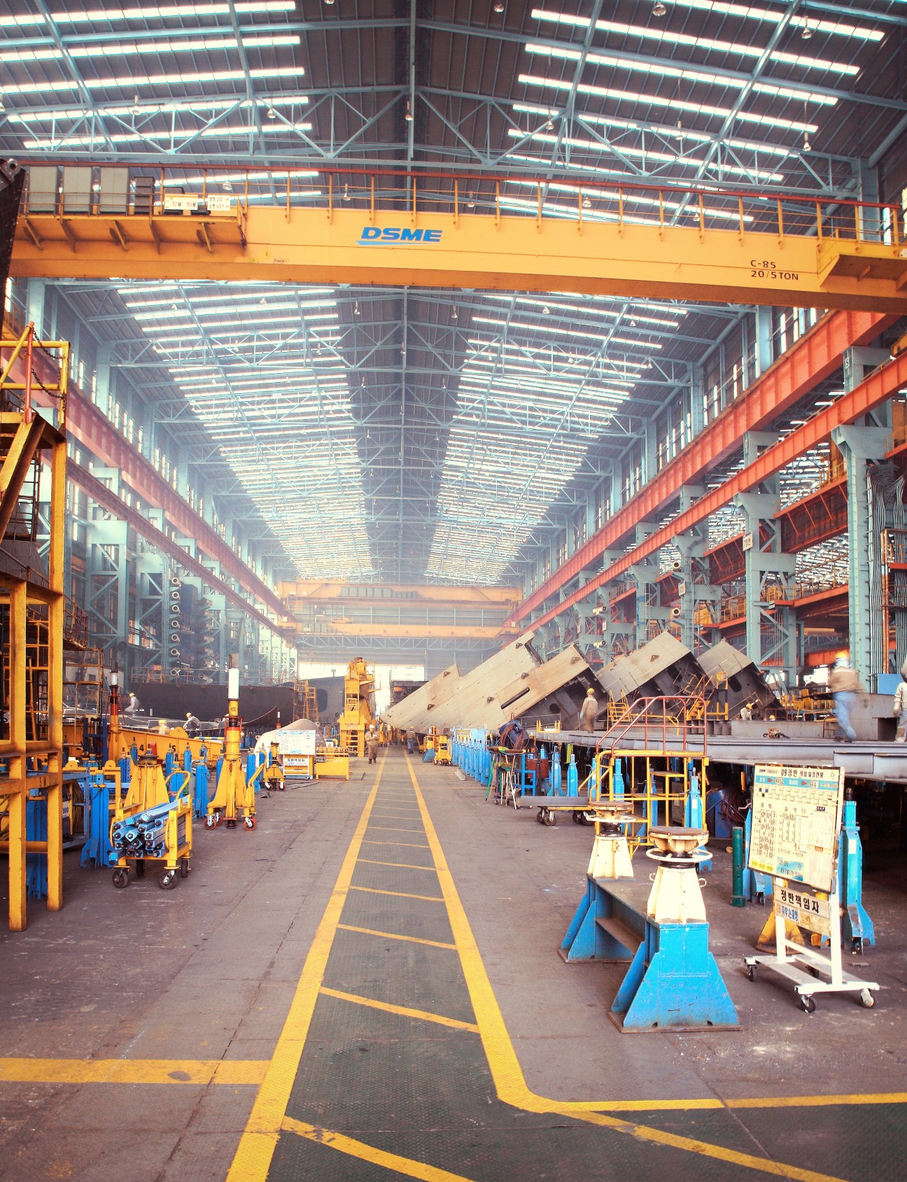 Interior of DSME assembly shop with yellow overhead cranes and blue equipment along yellow floor markings.