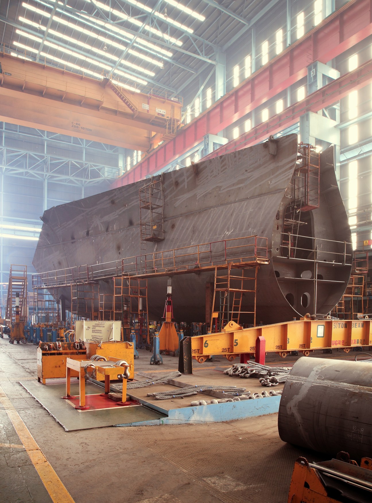 Industrial shipbuilding assembly shop with massive steel ship hull under construction, overhead cranes, and orange work equipment.