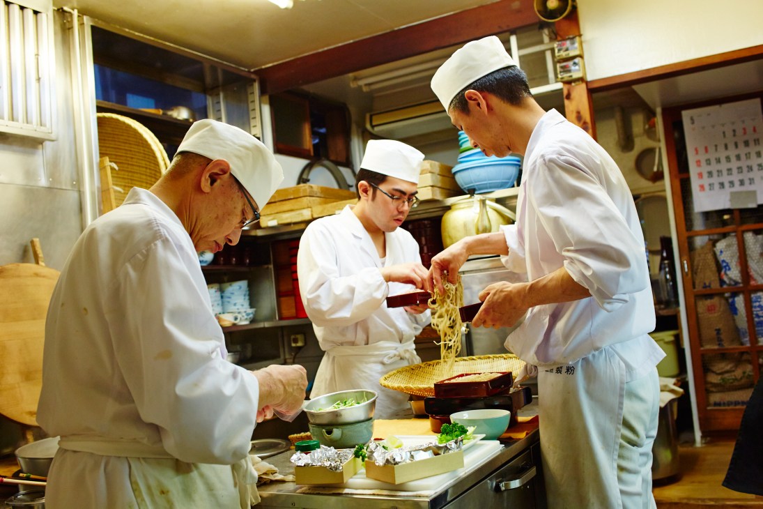 Three chefs in white uniforms and head coverings prepare soba noodles at a low table in a traditional Japanese kitchen.