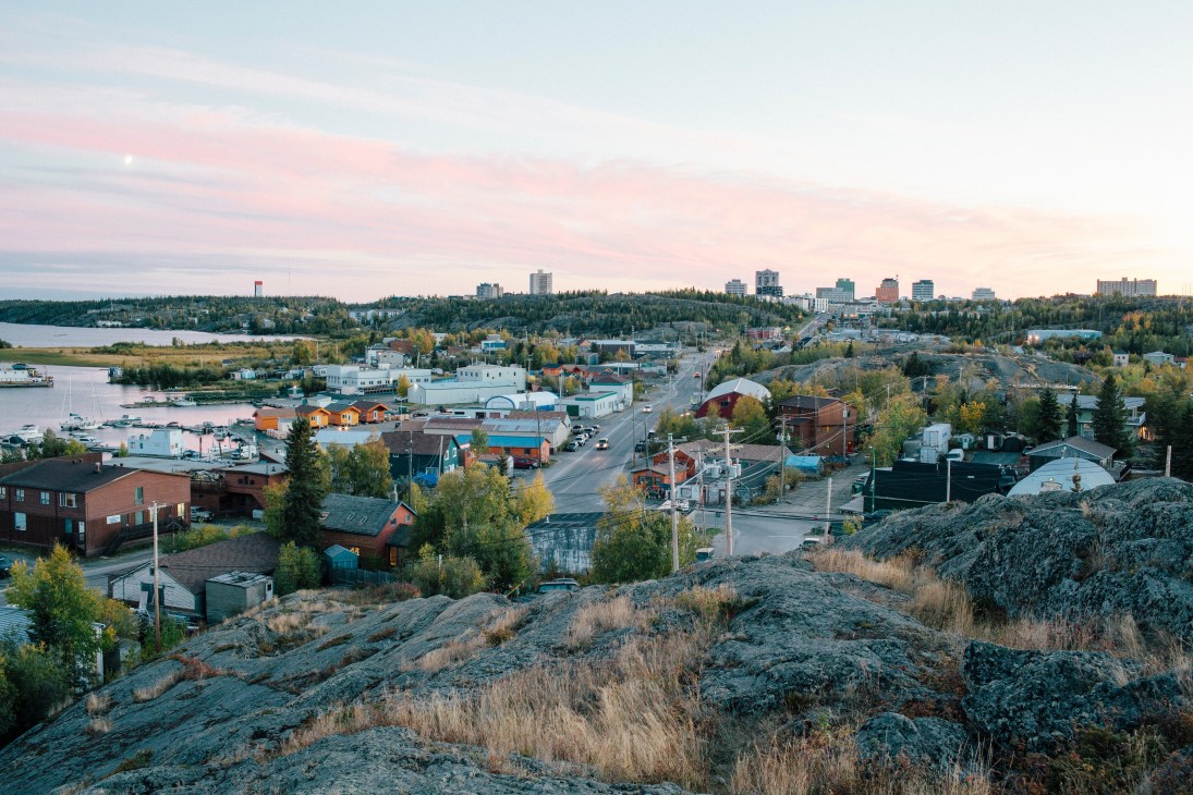 Aerial view of Yellowknife cityscape with buildings, water, forests and rocky terrain at dusk.