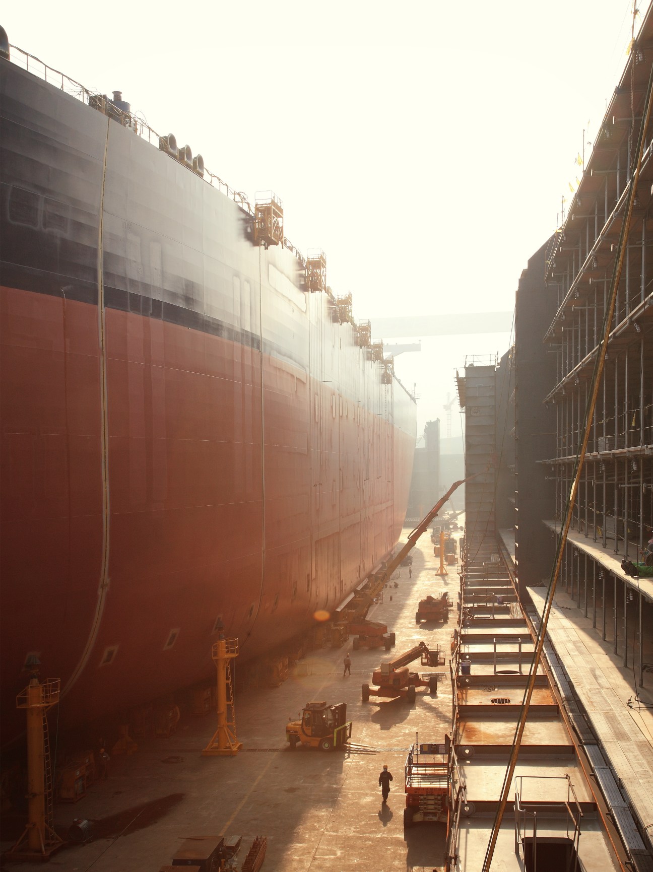Large red oil tanker under construction in a massive shipyard dock with industrial structures and workers visible.