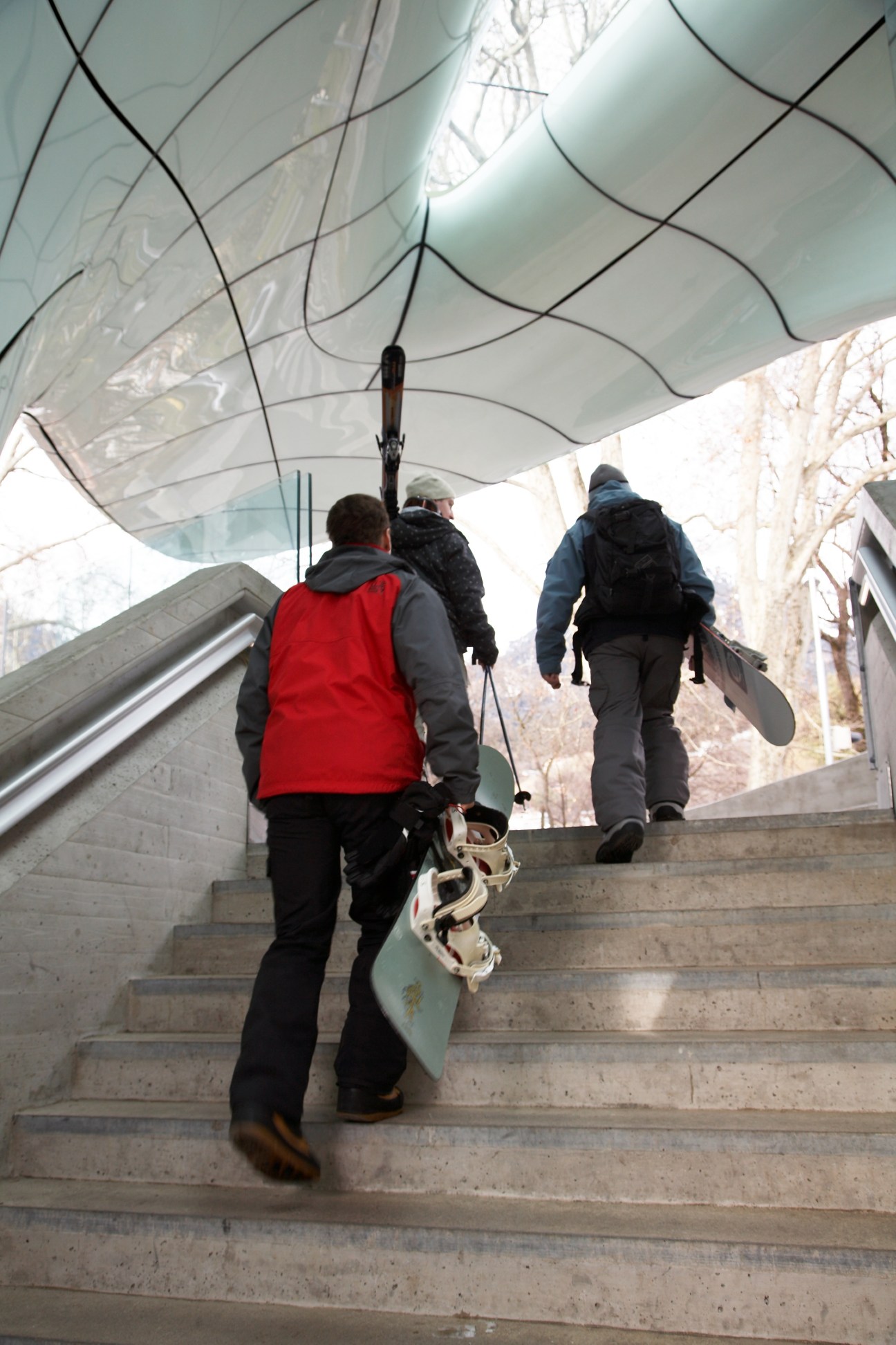Students ascending stairs under a modern curved canopy at Nordkettenbahn Congress station, carrying snowboards.