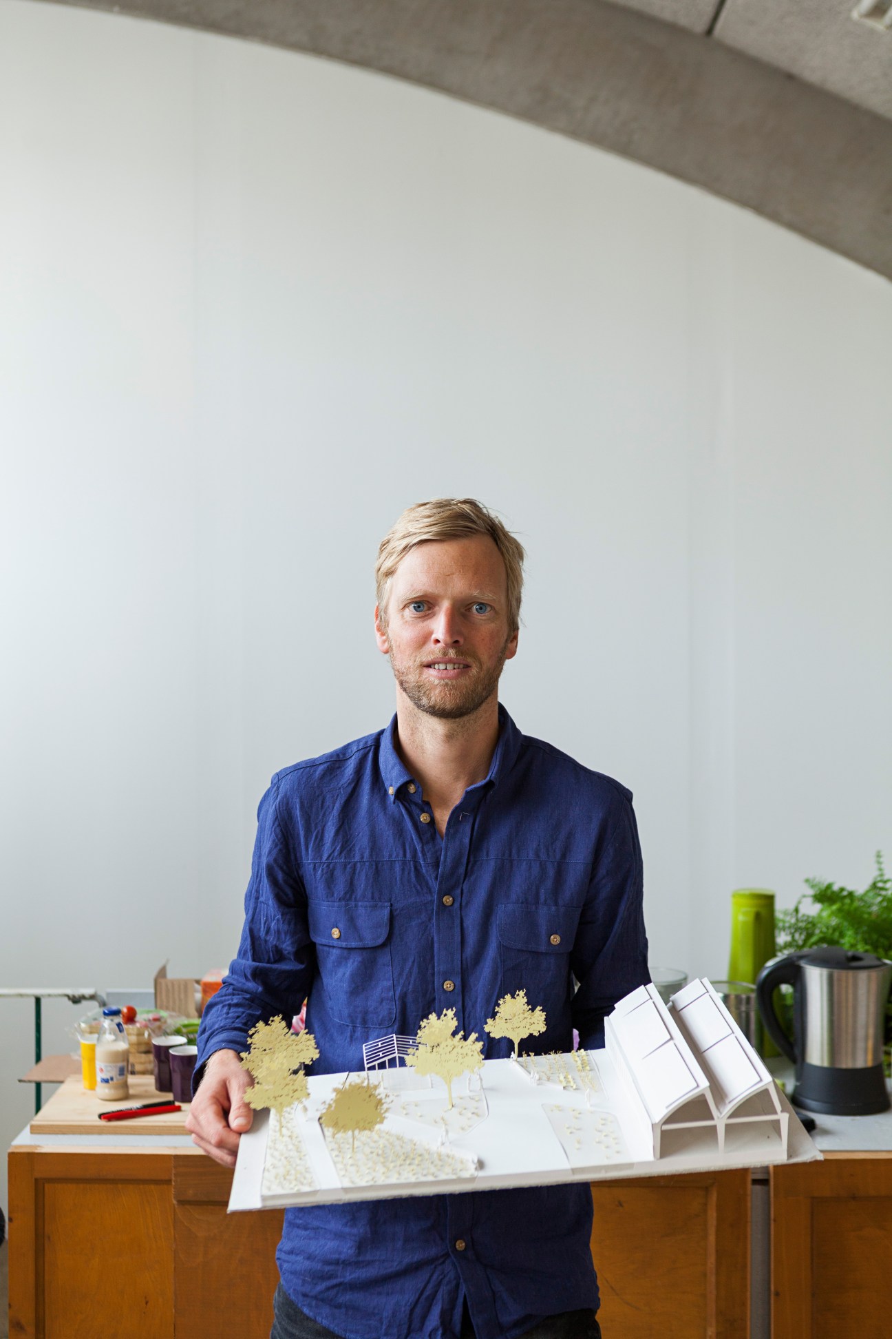 Gerwin de Vries, landscape architect and co-founder of Lint, stands in his studio holding architectural maquettes with a sloping concrete roof visible above.