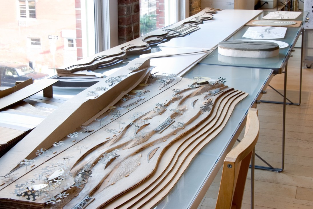 Architecture models and landscape design plans displayed on a table in an office with windows.