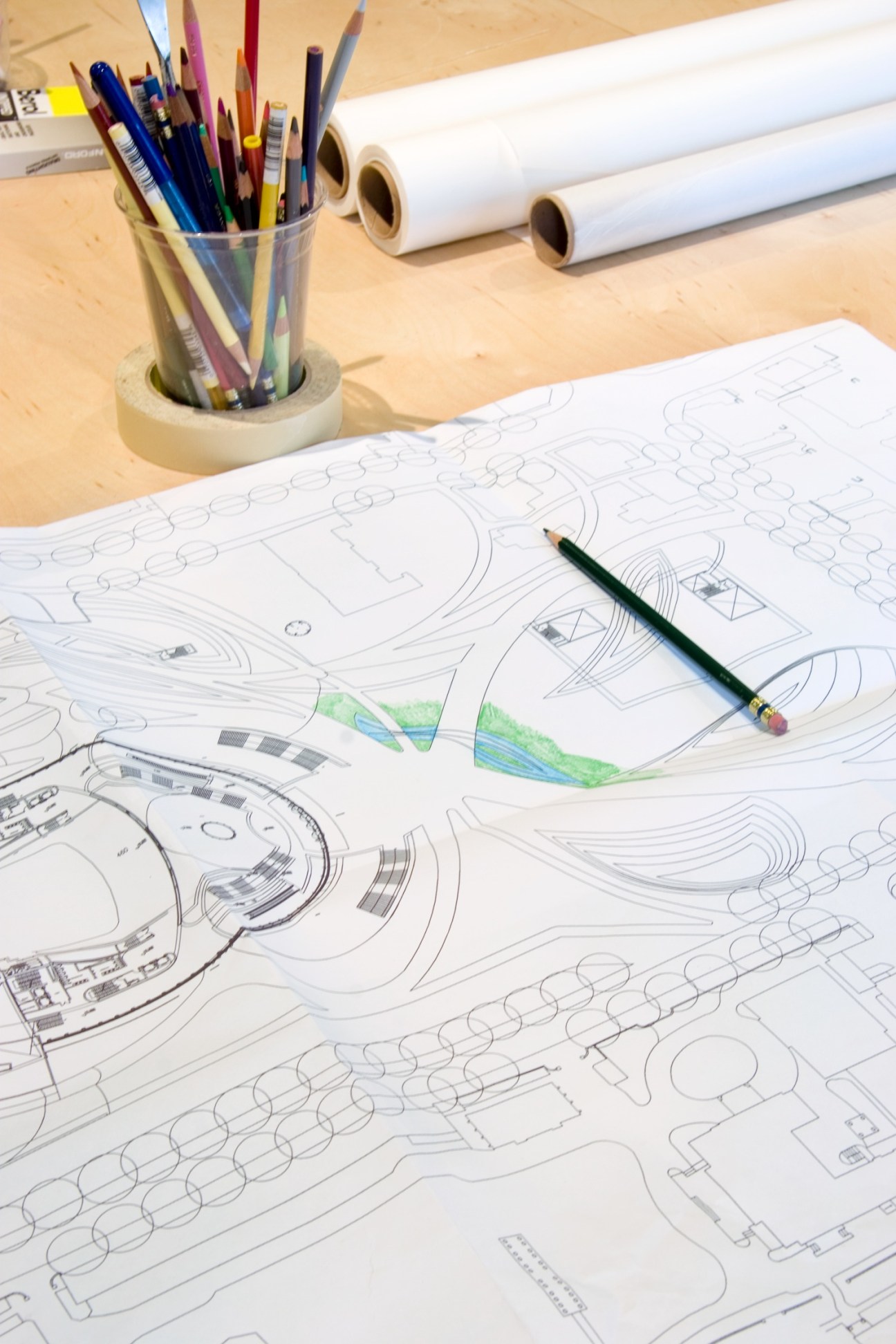 Architectural plans with sketches and green watercolor details on a wooden desk with colored pencils and design tools.