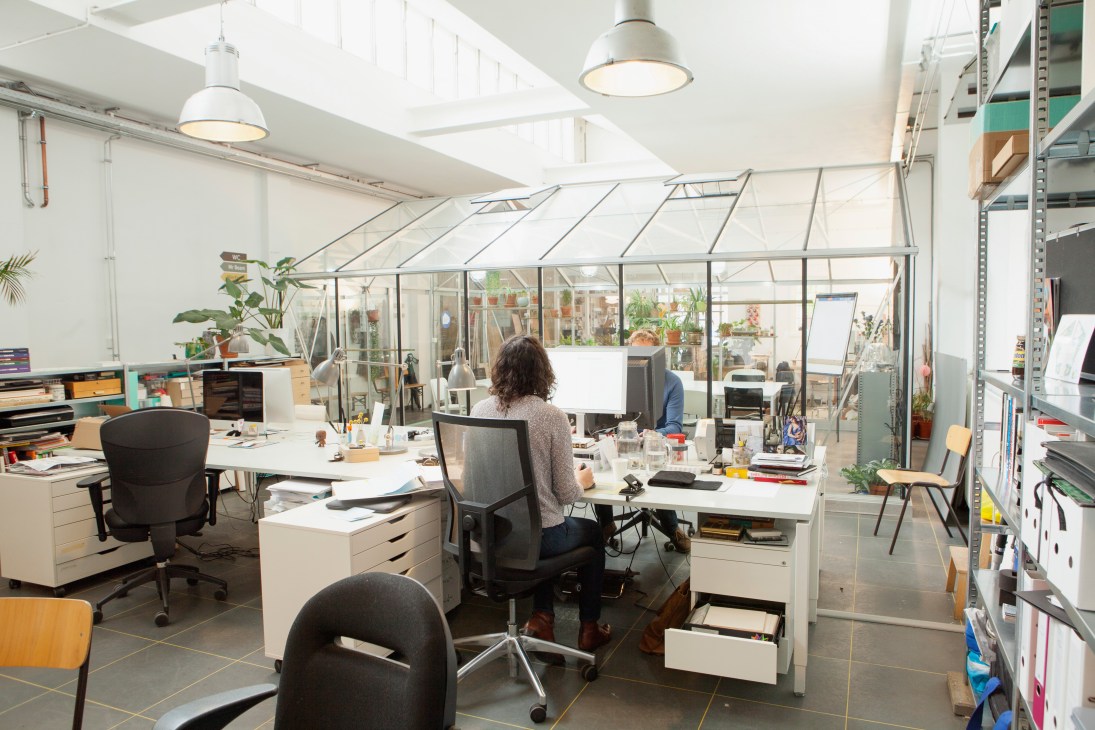 Open-plan shared workspace with white desks, workers, and a glass-walled greenhouse structure with plants