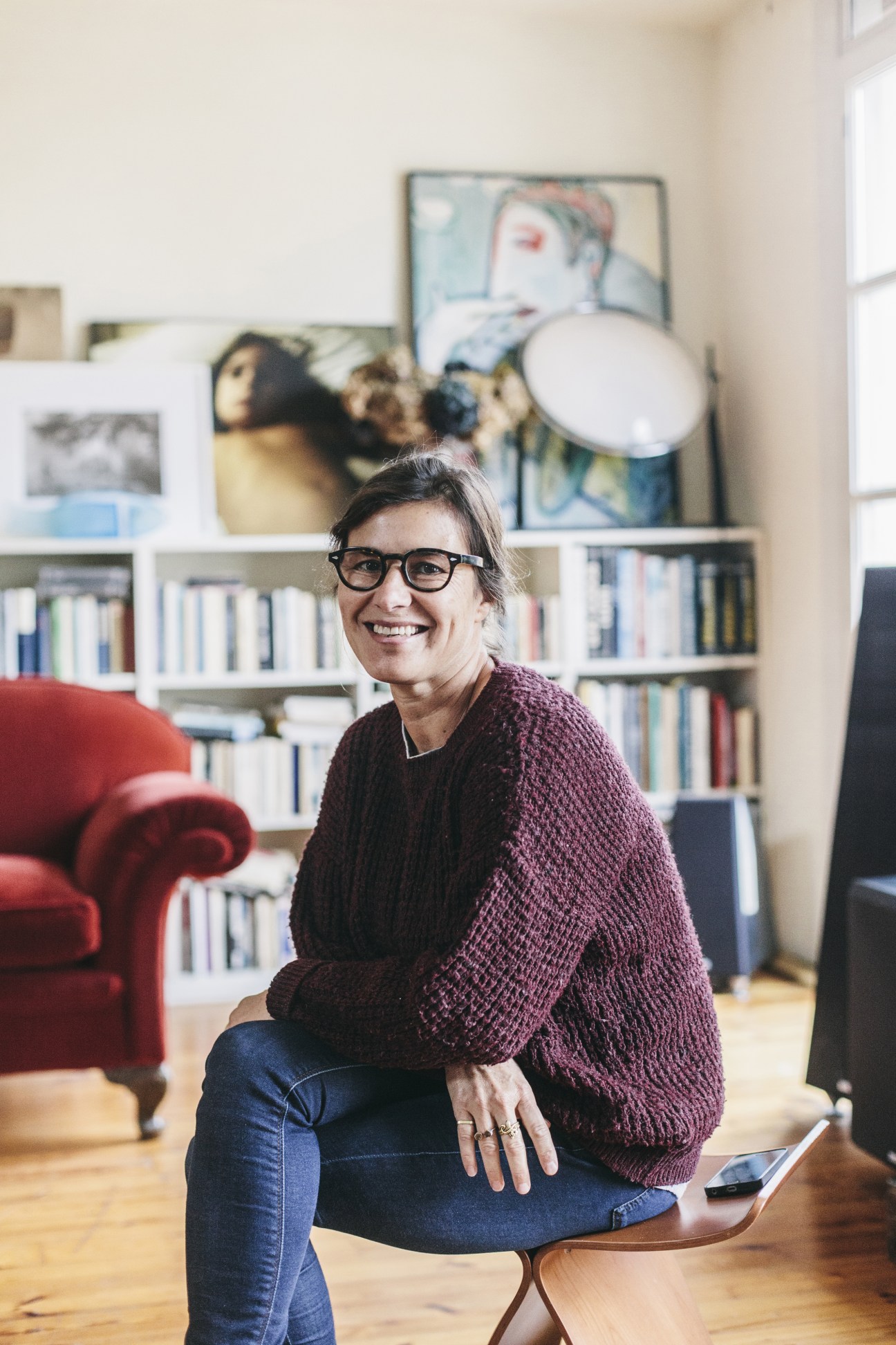 Woman in glasses and burgundy sweater smiling in a modern living room with bookshelves and artwork.