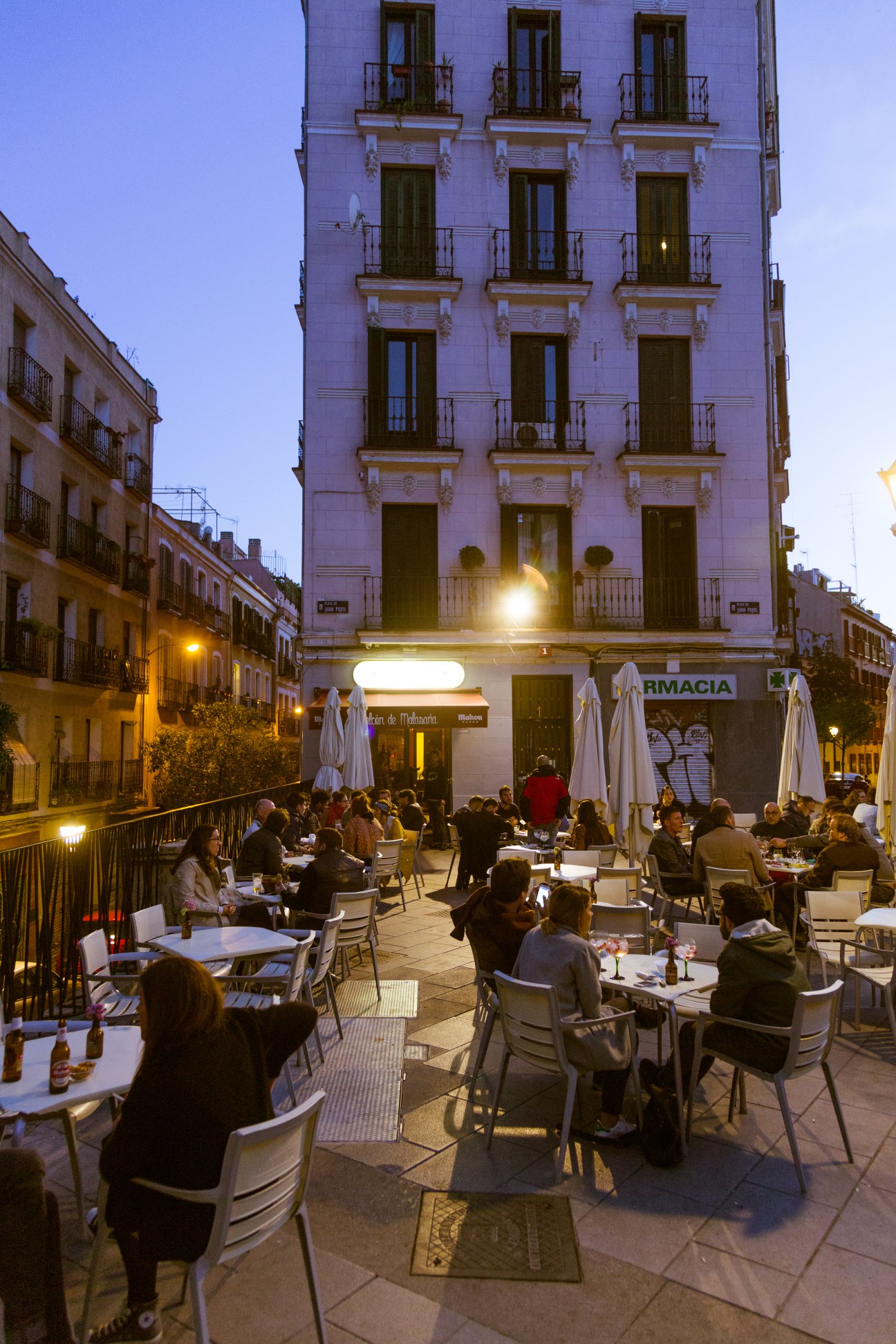 Evening diners seated at outdoor tables in Plaza Juan Pujol, Malasaña, with neoclassical buildings and white umbrellas.