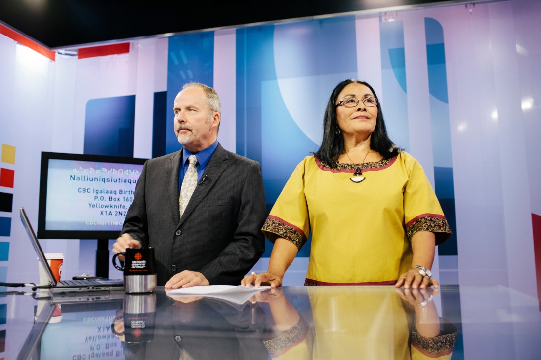 Two news anchors sit at a broadcast desk in a TV studio with blue geometric set design behind them.