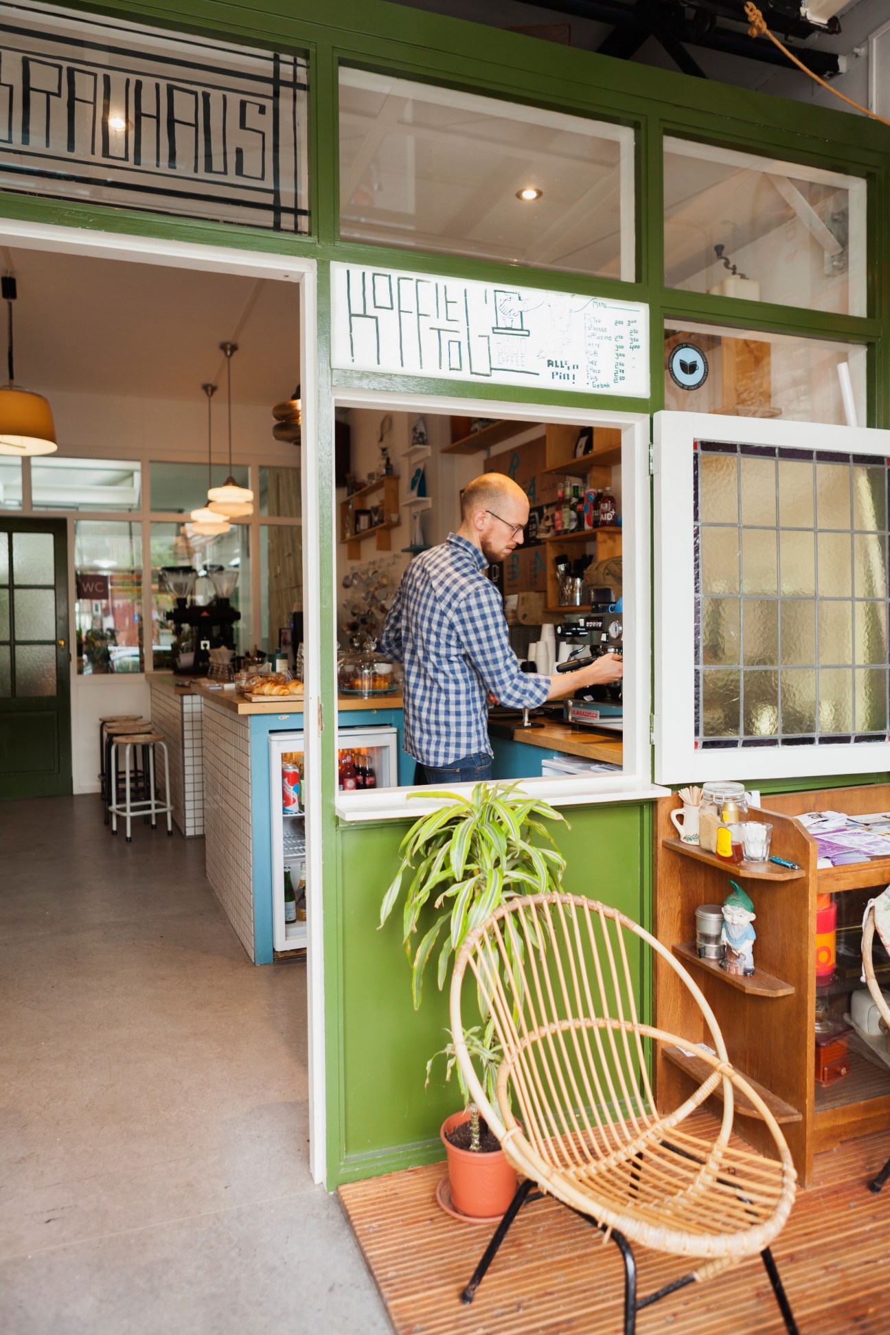 Man working at counter of Koffie Leute Brauhaus café with green storefront and potted plant outside.